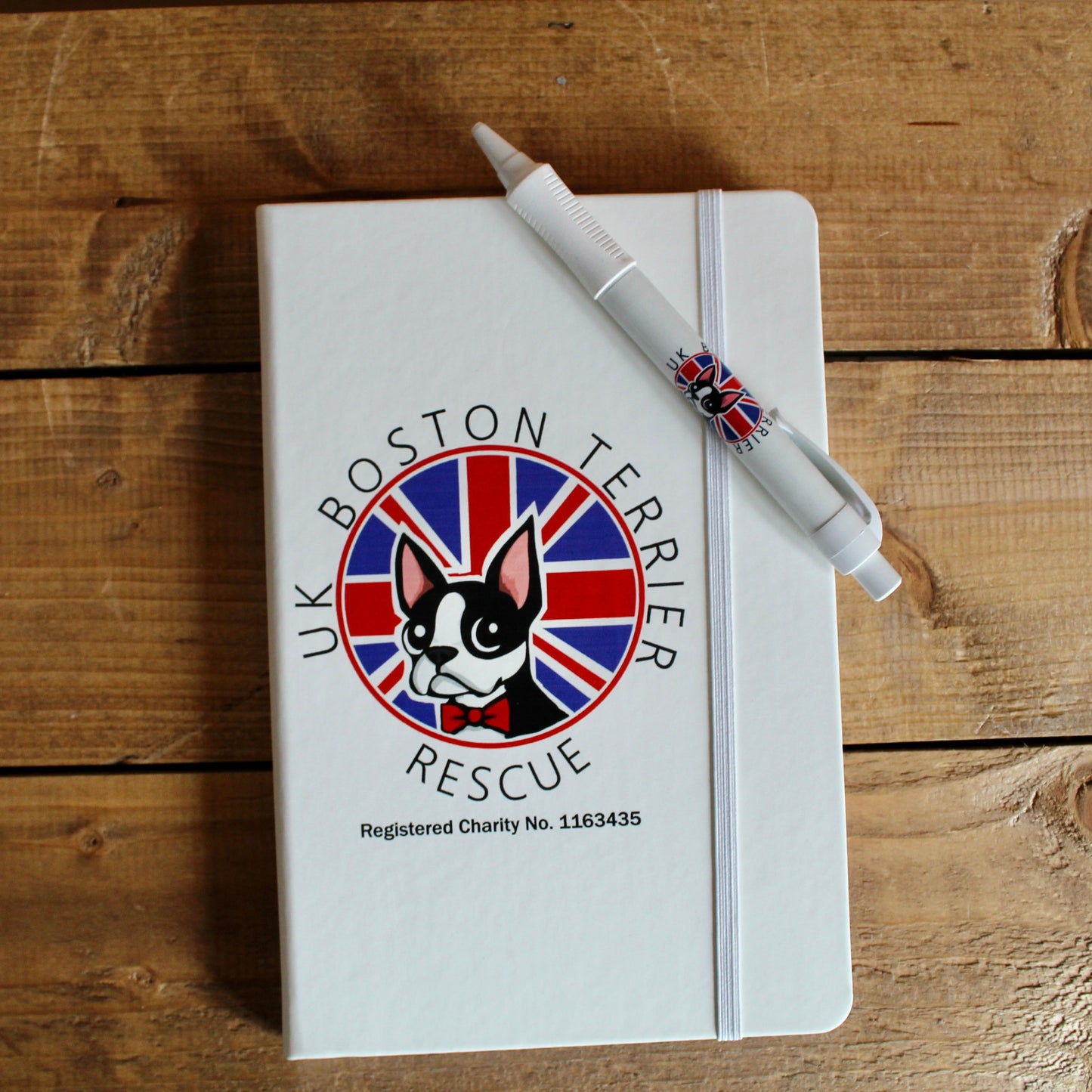 A white A5 size hardcover notebook with a pen placed on it. The notebook has a printed design featuring a dog and text related to the UK Boston Terrier Rescue.