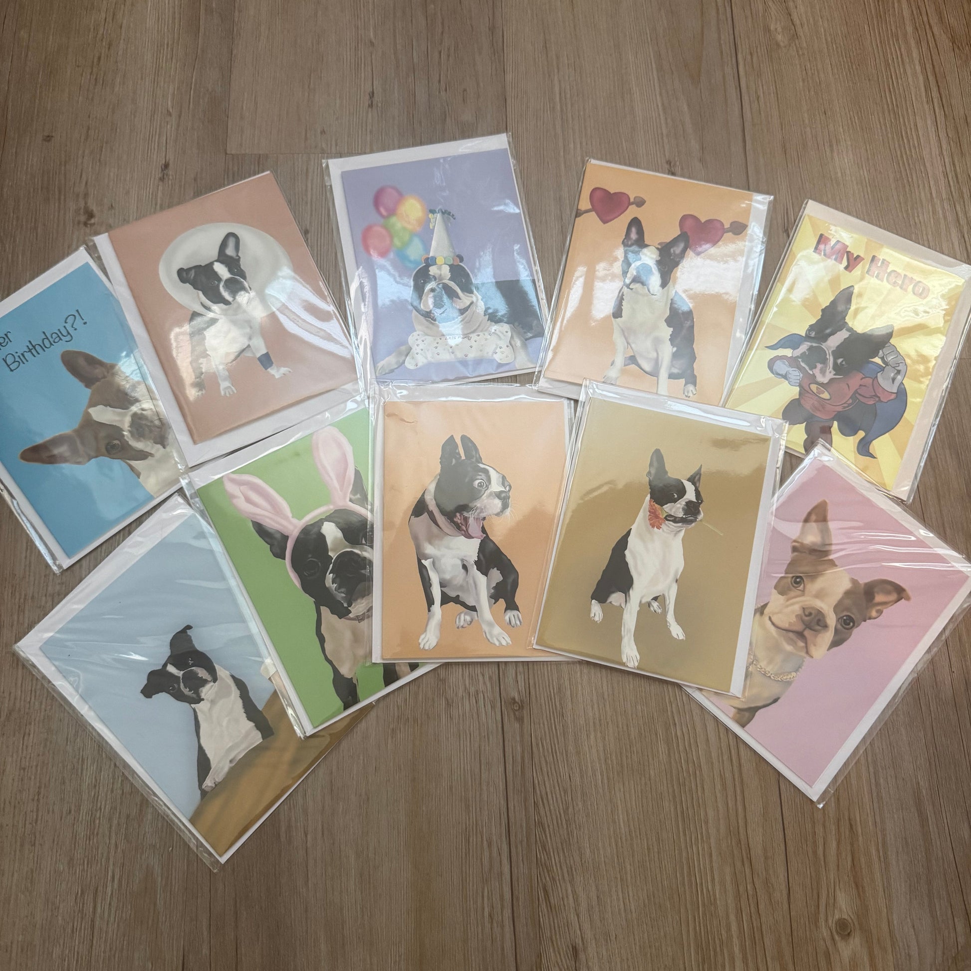 A set of dog-themed greeting cards arranged in a loose circle on a wooden floor. Each card features a different colored background with an illustration of a French Bulldog in various poses.