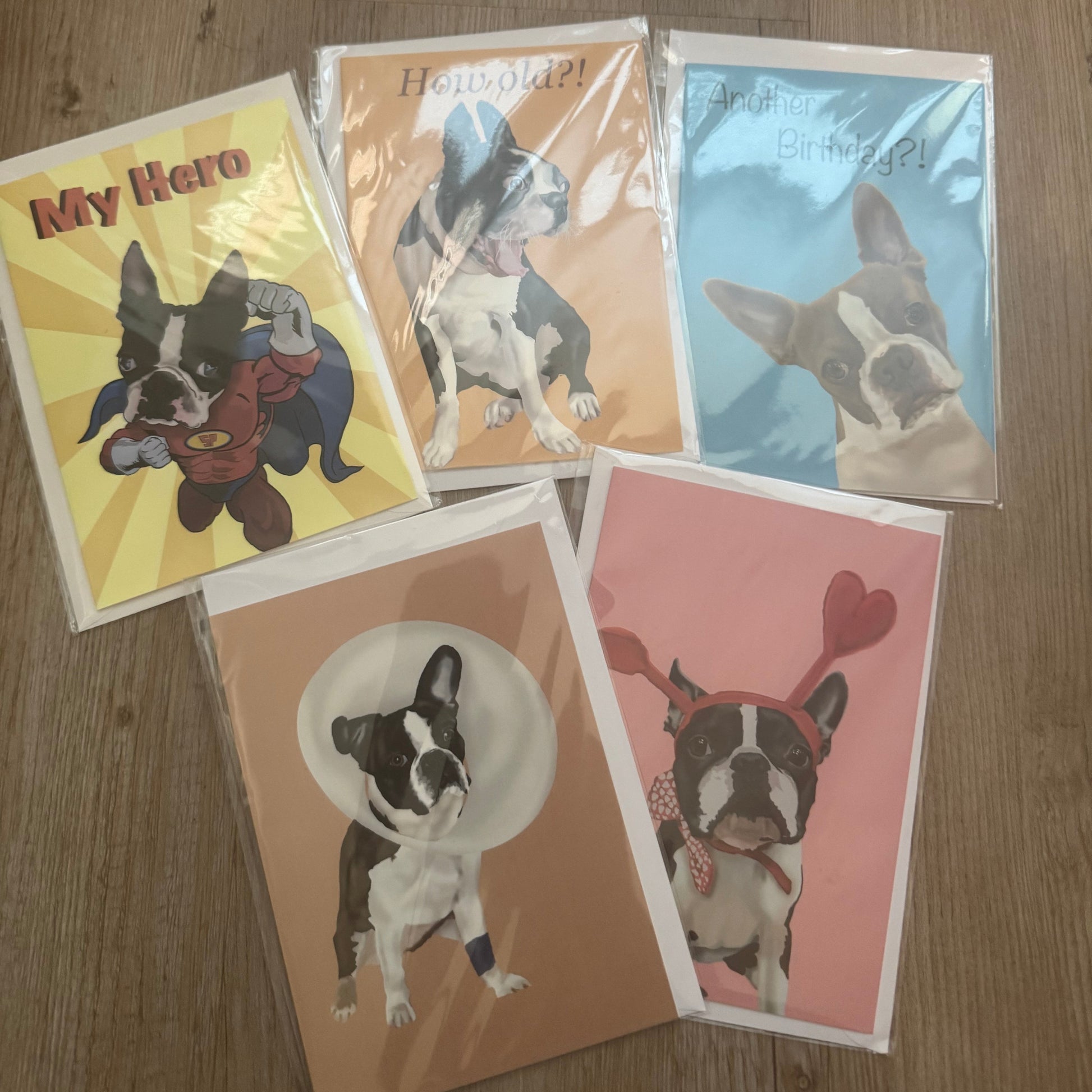 Five printed cards featuring illustrated Boston Terrier dogs in different poses and colors, arranged on a wood floor. The cards have white borders and are photographed from above.
