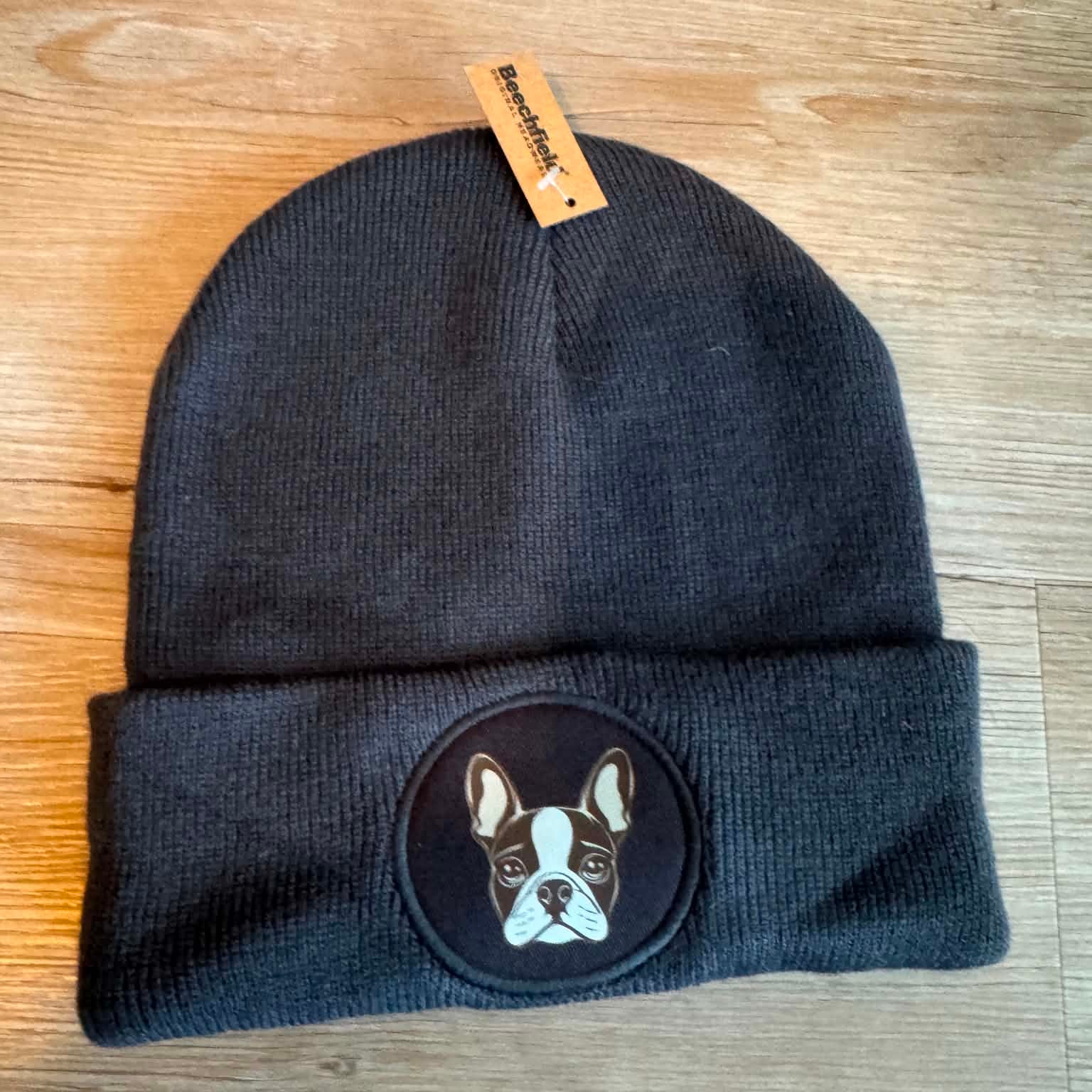 A dark charcoal gray knit beanie hat with a folded cuff, featuring a circular embroidered patch of a French Bulldog on the front. A small tan paper tag is attached at the top.