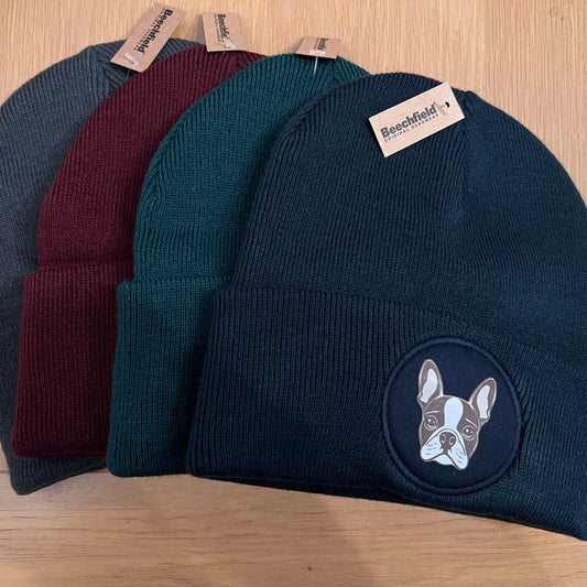 A collection of beanie hats in gray, red, and blue colors, displayed on a wooden surface, with a patch on the blue beanie depicting a dog.