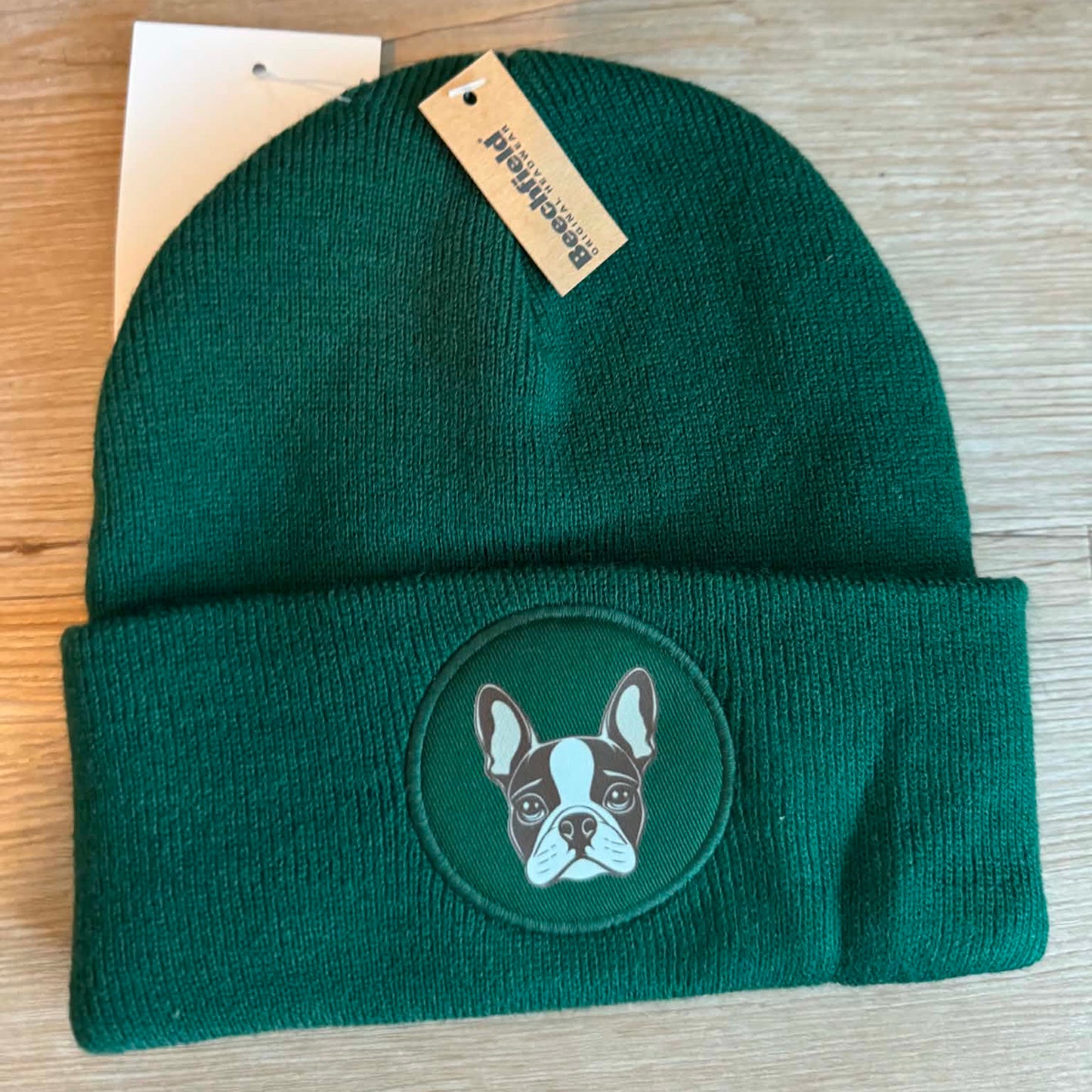A dark green knit beanie with a folded cuff featuring a circular embroidered patch of a Boston Terrier dog. A small paper hangtag is attached near the top, and the hat is laid flat on a light wood surface.