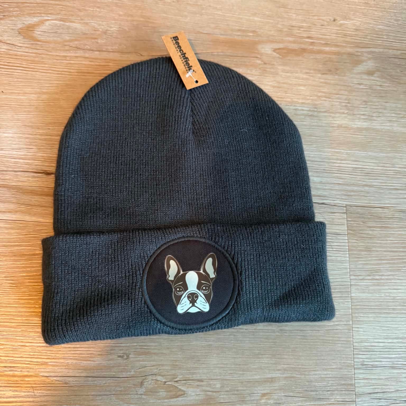 A dark charcoal gray knit beanie hat with a folded cuff, featuring a circular embroidered patch of a French Bulldog face on the front. A small tan brand tag is attached at the top.