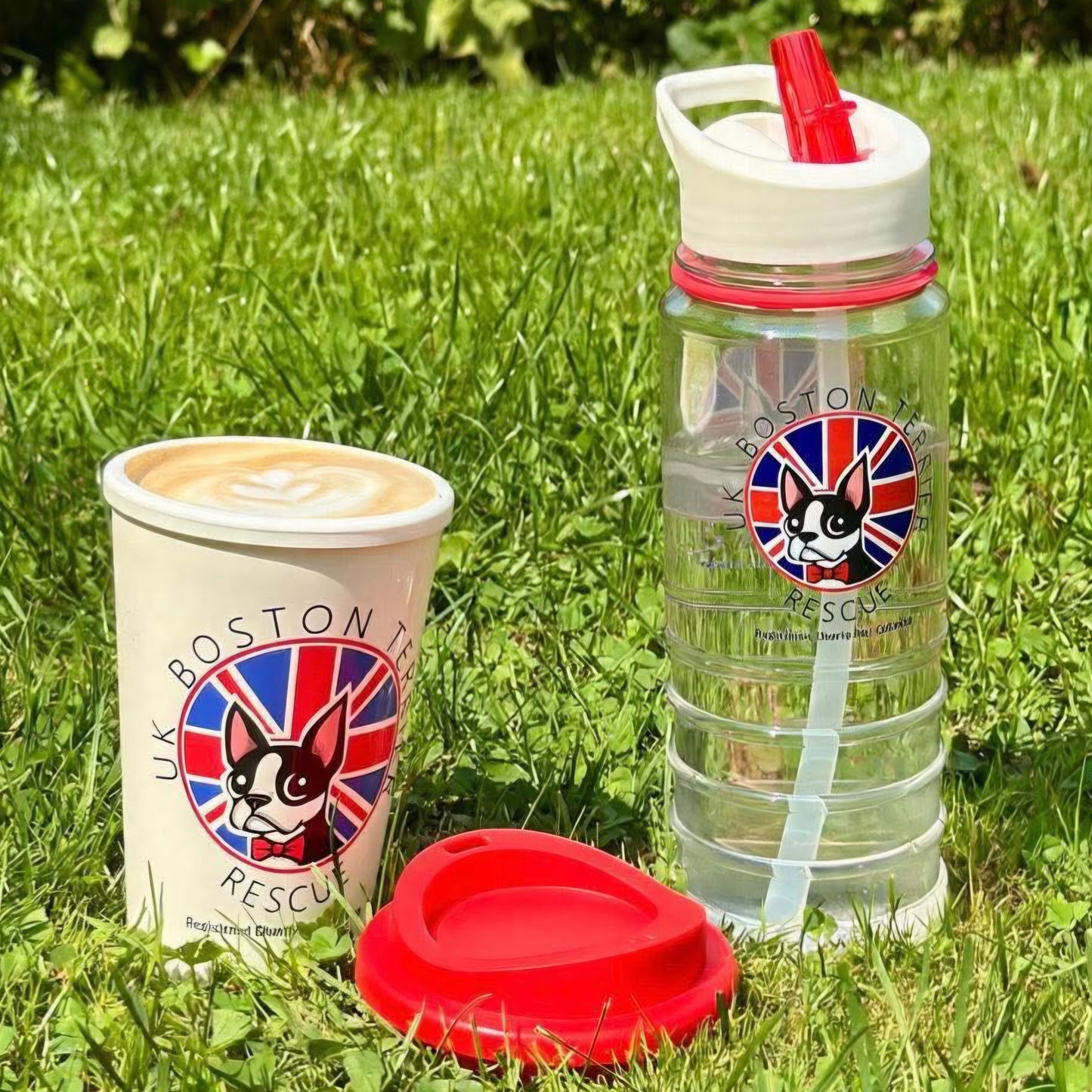 A clear plastic water bottle with a white lid and red straw, alongside a white coffee cup with a printed Boston Tea Rescue logo, and a red lid, all placed on green grass outdoors.