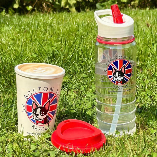 A clear plastic water bottle with a white lid and red straw, alongside a white coffee cup with a printed Boston Tea Rescue logo, and a red lid, all placed on green grass outdoors.