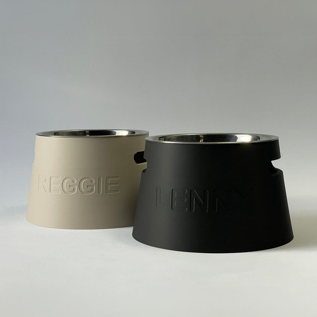 Two pet food bowls on a light gray studio background. The front bowl is matte black with an embossed 'ILIO' logo and a stainless steel rim.