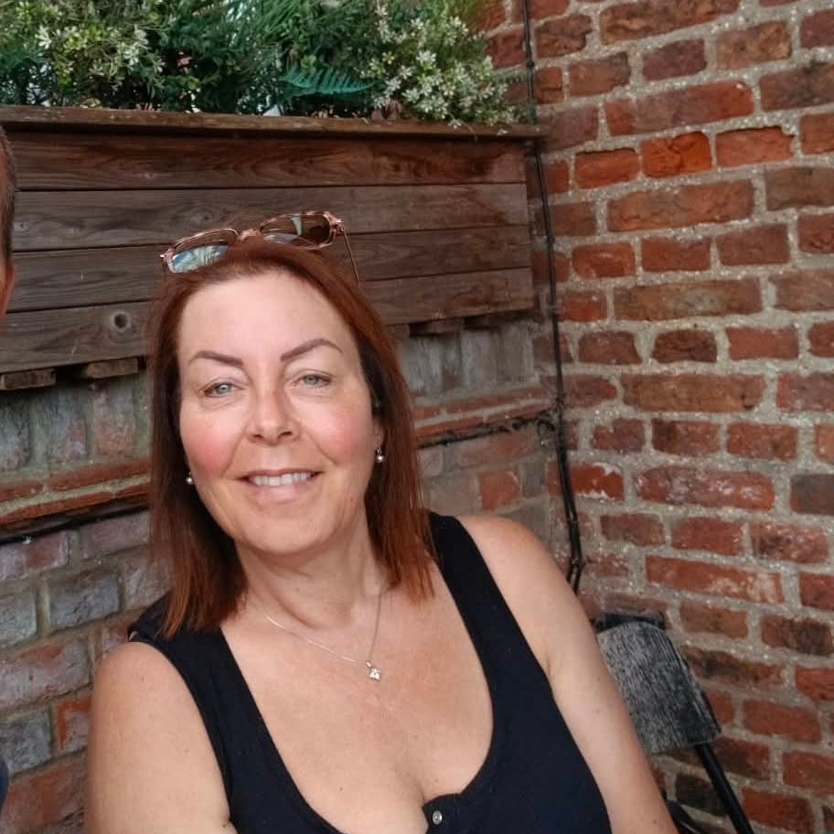 A smiling woman with brown hair and glasses on her head sits in front of a red brick wall and wooden planter, wearing a black sleeveless top.
